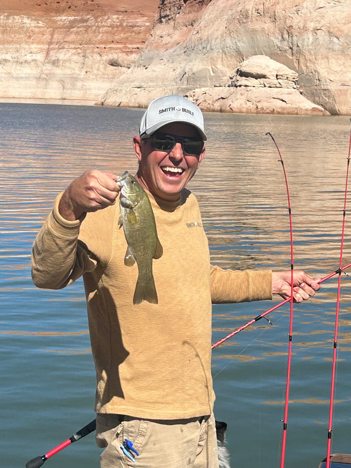 Tyler Smith, owner of Smith Built, in a Smith Built cap and sunglasses on a boat, holding up a smallmouth bass, with red sandstone cliffs and open water behind him