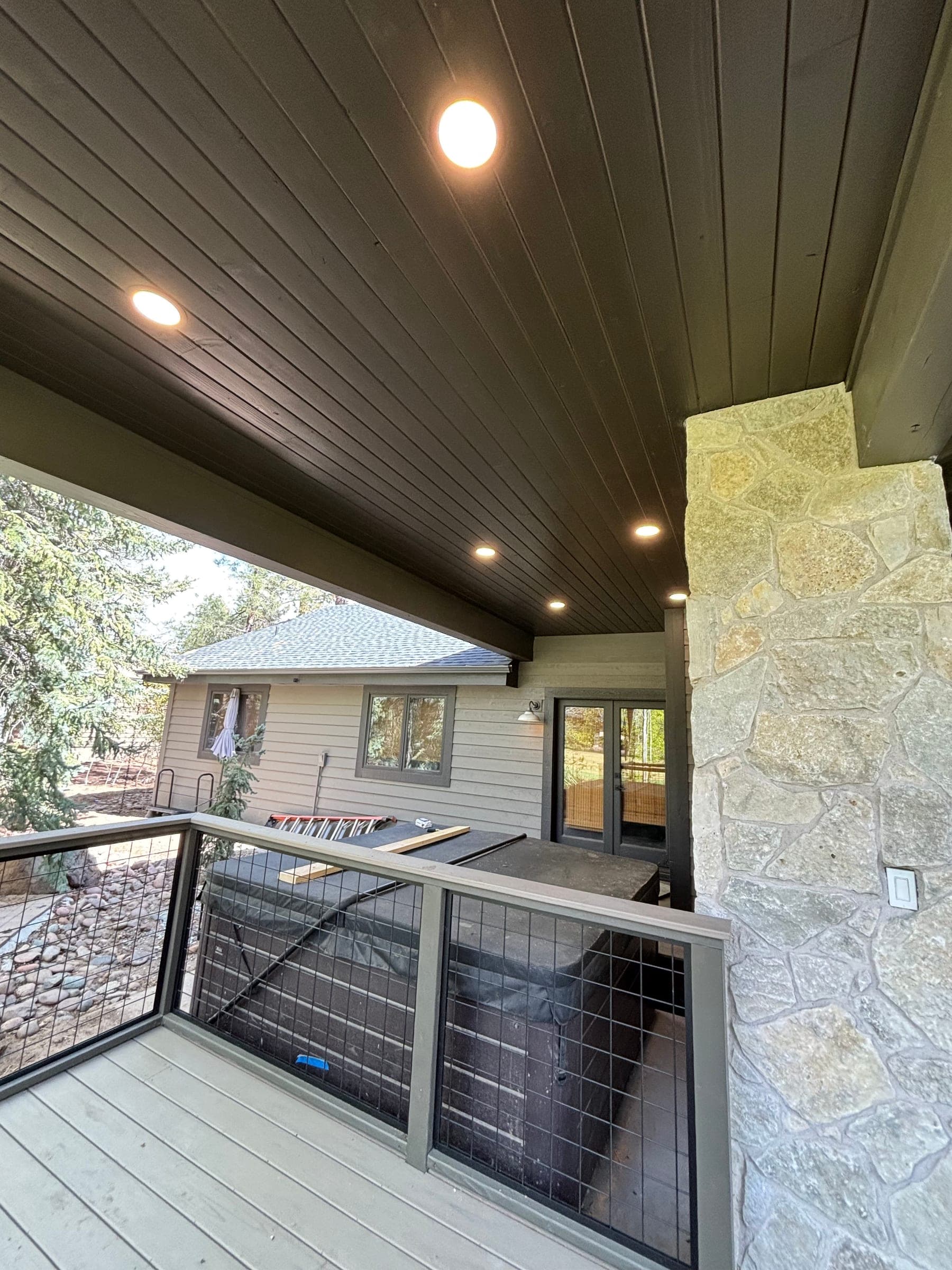 Covered deck with dark stained soffit, a stone-clad column and welded wire-mesh panel railing, with pines beyond