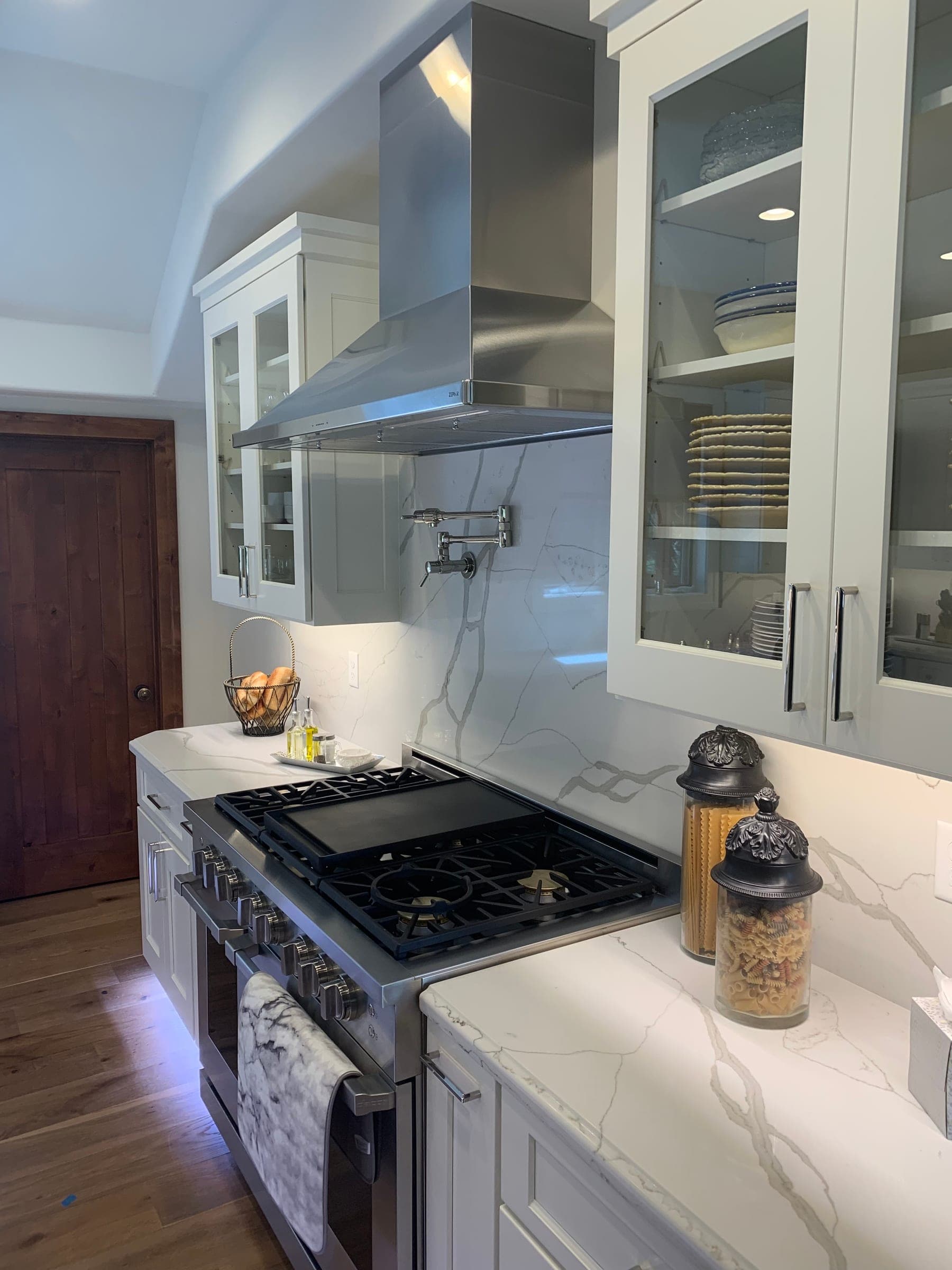 Kitchen range wall with a stainless hood, slab backsplash, wall-mounted pot filler and glass-front upper cabinets