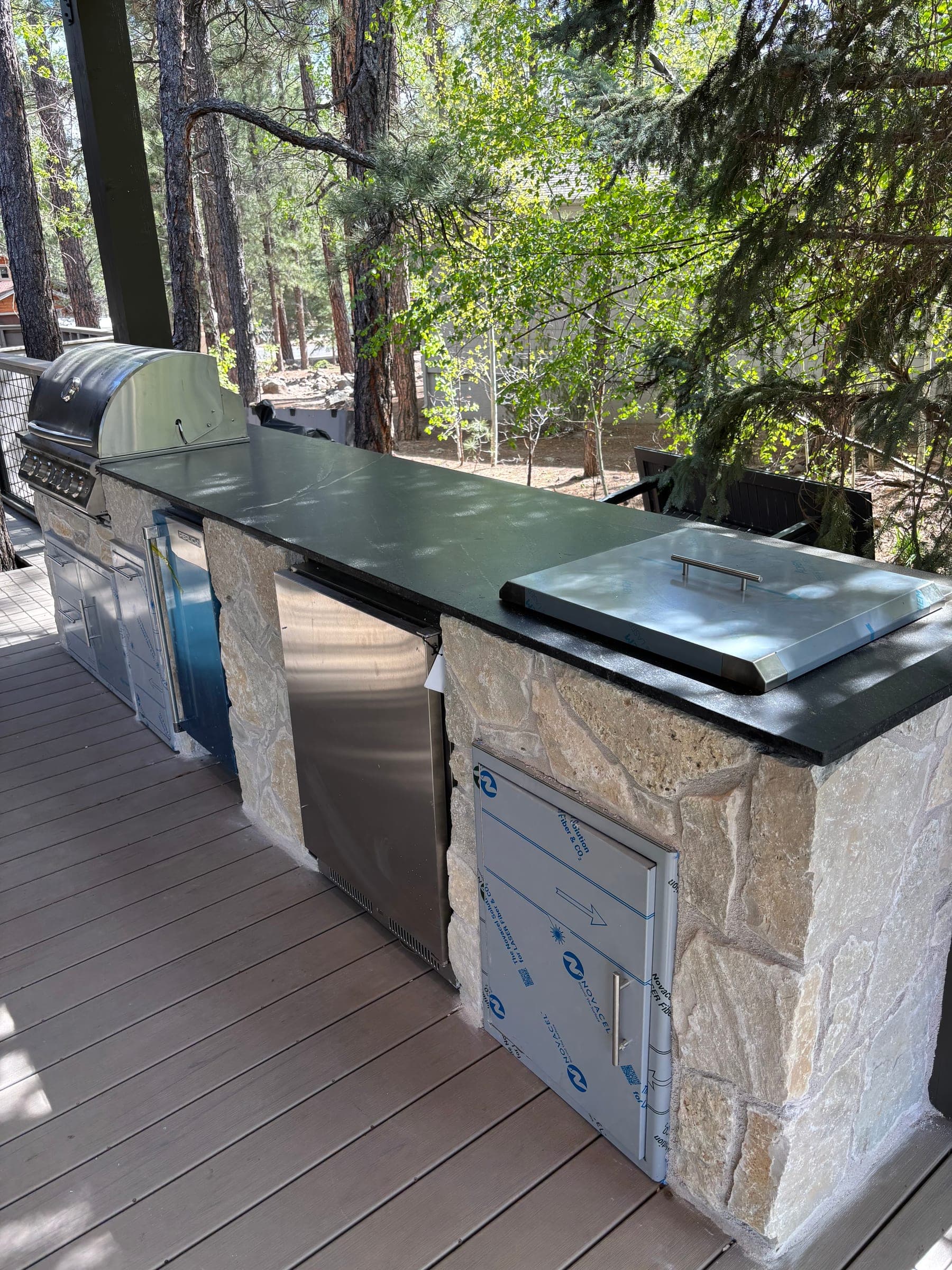 Stone-faced outdoor kitchen counter with dark stone top, stainless grill and access doors still covered in protective film