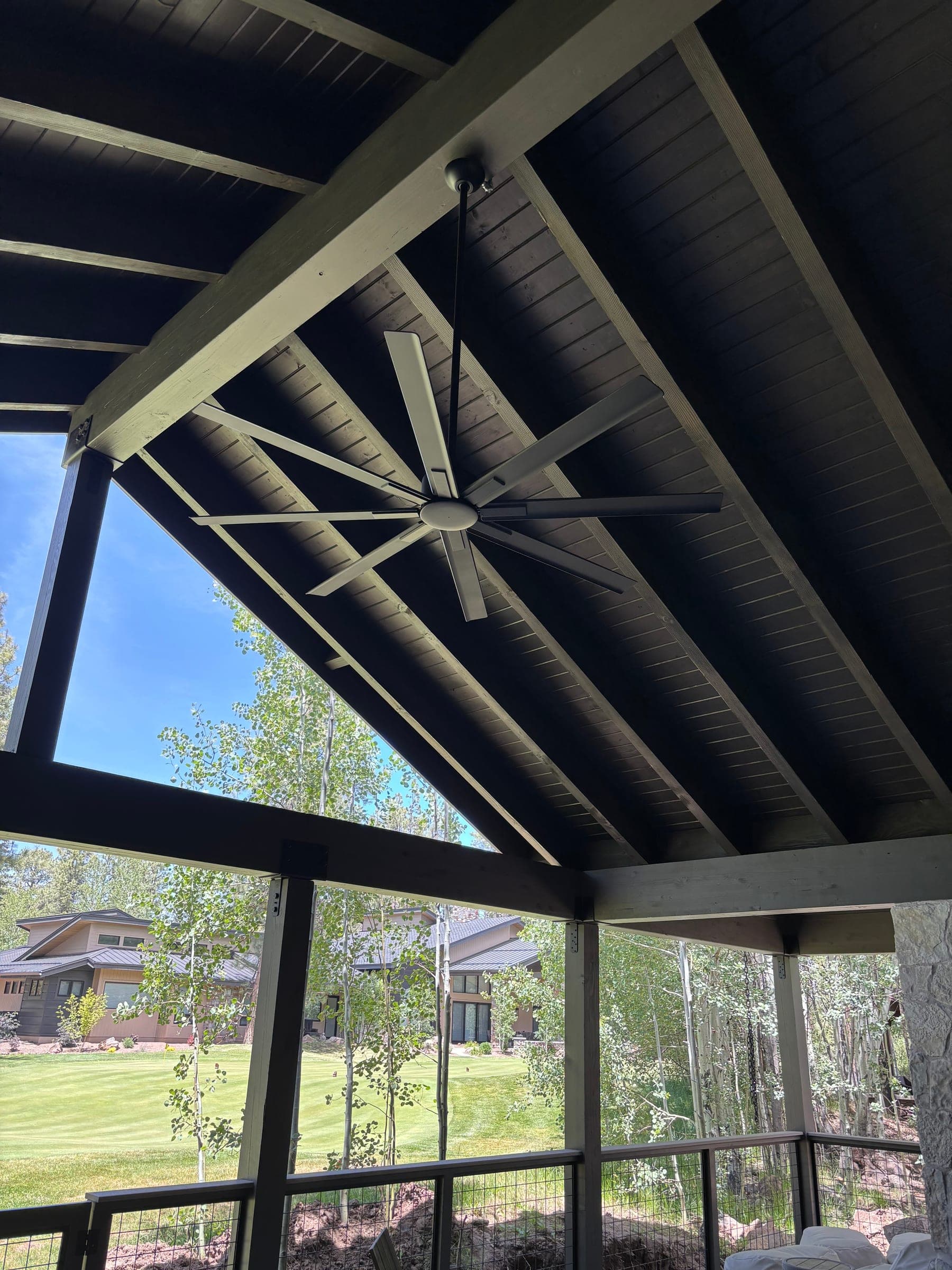 Covered patio with light-painted exposed rafters over a dark tongue-and-groove ceiling and a large-span ceiling fan, aspens beyond